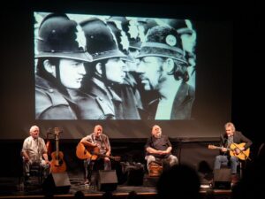 Photo of four musicians in Pitmen Poets on stage with visual screen projection of miners and police behind